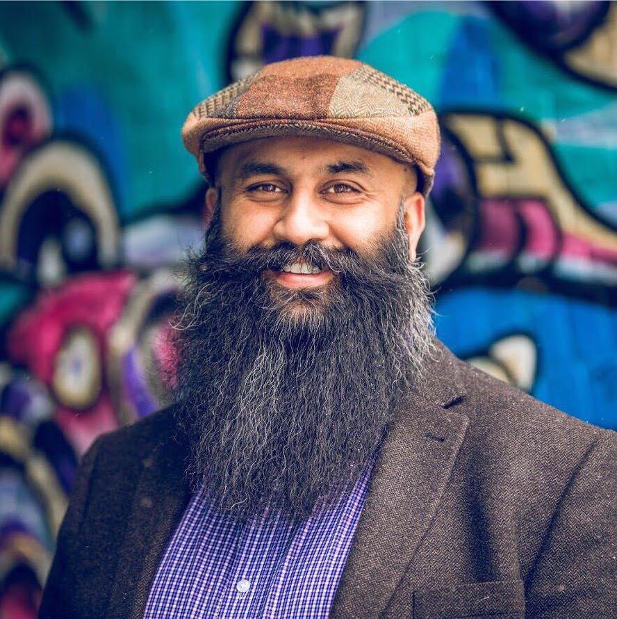 Hasan smiles in front of a tasteful graffiti wall. He is wearing a brown cap, a grey blazer, a blue/white checkered shirt, and a glorious beard.